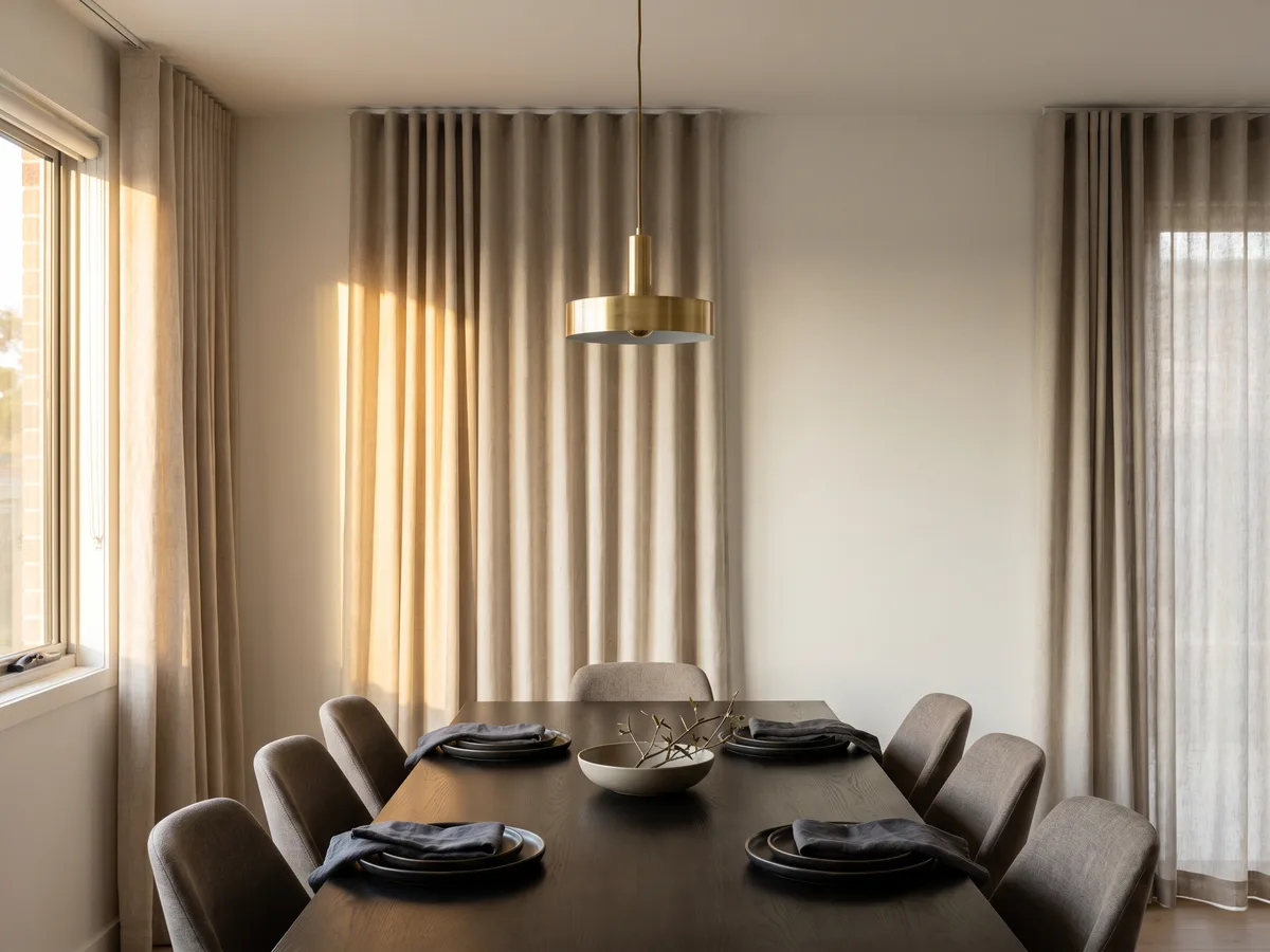 Dining room with floor-length oatmeal linen curtains on an S-fold track, late sun catching one fold beneath a brass pendant